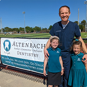 Jacksonville dentist with his daughters next to poster for Altenbach Dentistry at local sports game