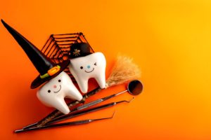 Two toy teeth dressed up in Halloween hats sitting on an orange background with dental instruments and a witches broom